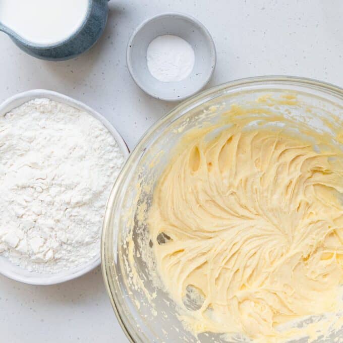 Tinned Plum Sponge Pudding A glass bowl of yellow cake batter sits next to a bowl of flour, a small dish of baking powder, and a jug of milk on a white countertop.
