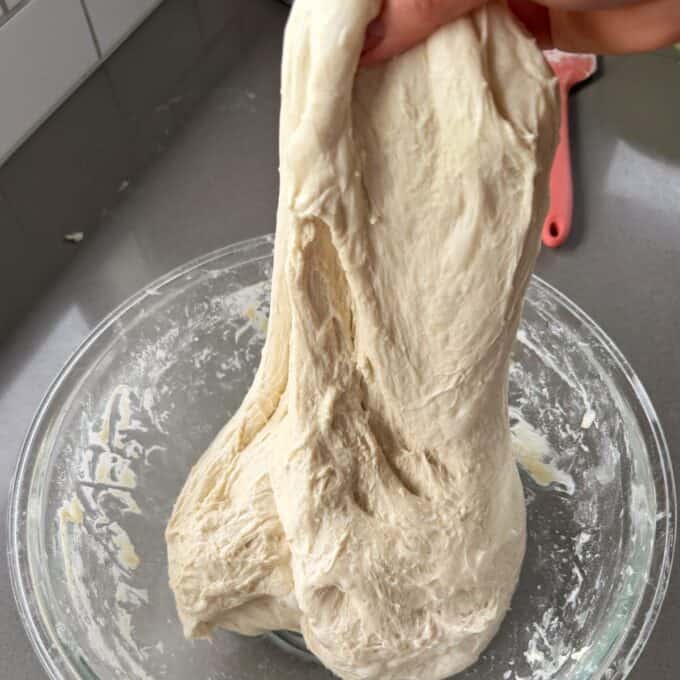 XL No-Knead Focaccia A hand stretches a portion of risen dough above a glass bowl on a gray countertop, with a pink spatula in the background.