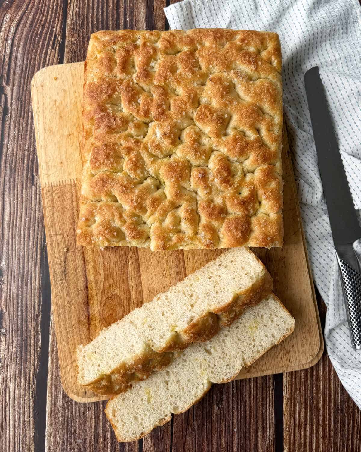 XL No-Knead Focaccia A square loaf of focaccia bread sits on a wooden cutting board, with two slices cut. A bread knife and a white cloth are beside the board.