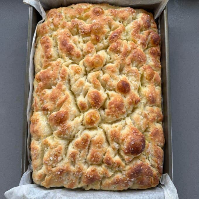 XL No-Knead Focaccia A rectangular pan of golden-brown focaccia bread with a dimpled surface, sprinkled with coarse salt, rests on parchment paper.