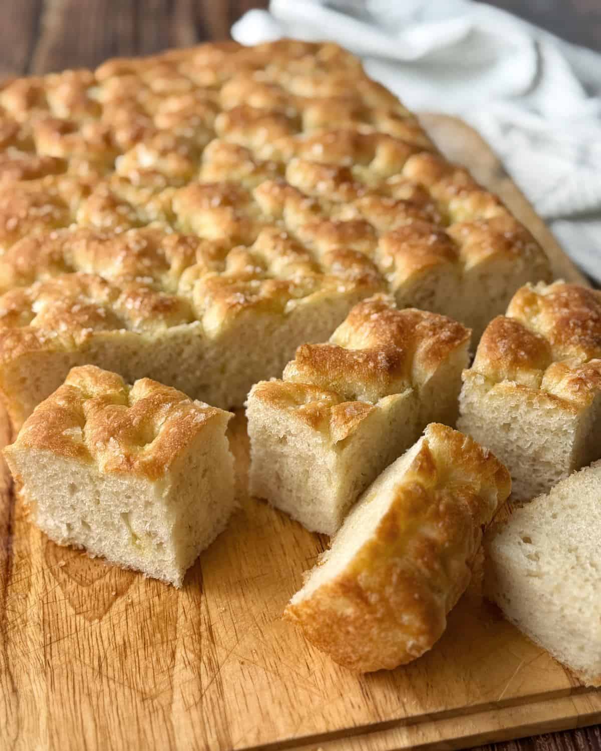 XL No-Knead Focaccia A rectangular loaf of focaccia bread on a wooden board, with several square pieces cut and arranged in front.