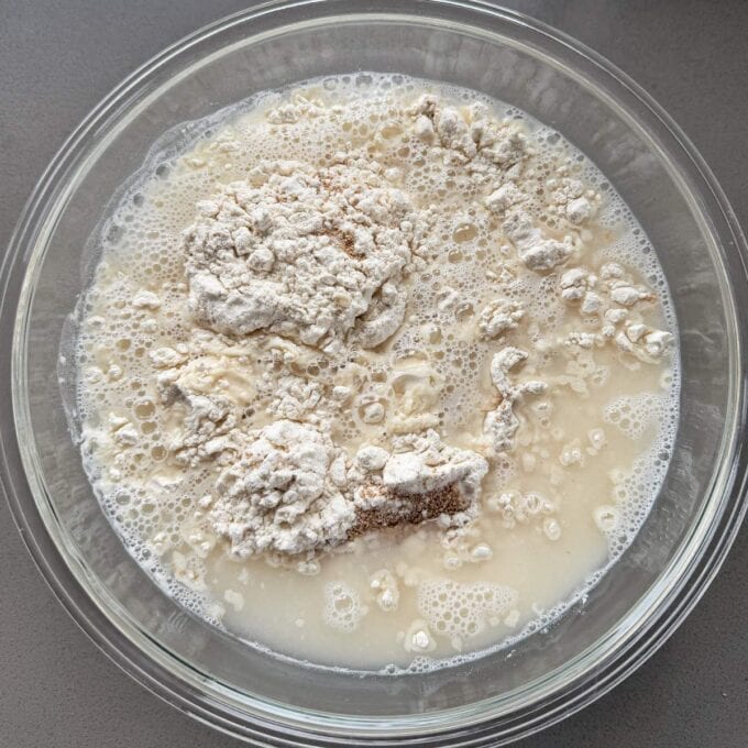 XL No-Knead Focaccia A glass bowl containing flour and liquid, possibly water or milk, before mixing, on a grey surface.