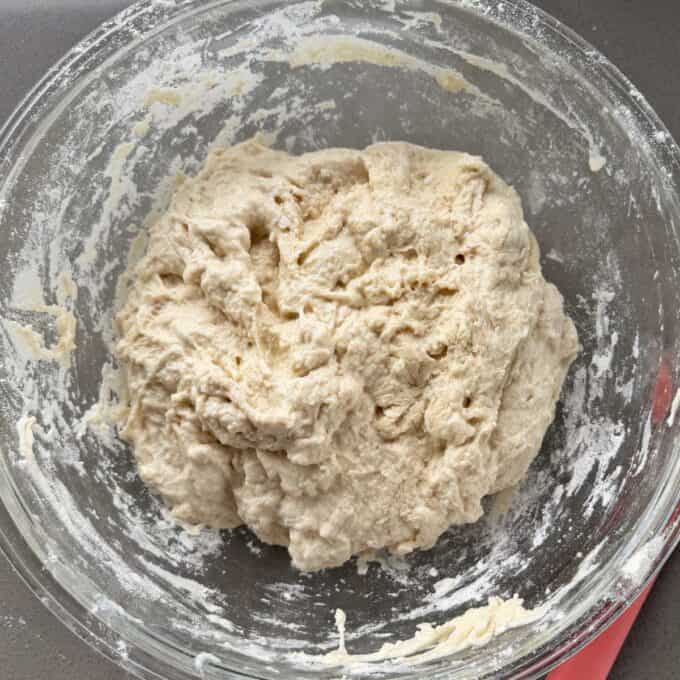 XL No-Knead Focaccia A glass bowl containing raw bread dough with some flour on the sides of the bowl, sitting on a grey countertop.