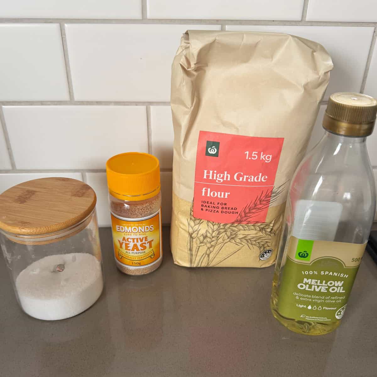 XL No-Knead Focaccia A jar of sugar, a container of active yeast, a 1.5 kg bag of high grade flour, and a bottle of mellow olive oil are arranged on a kitchen counter in front of a white tile wall.