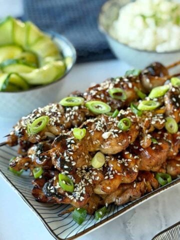 Grilled chicken skewers topped with sesame seeds and sliced green onions, served on a rectangular plate with cucumber salad and a bowl of rice in the background.