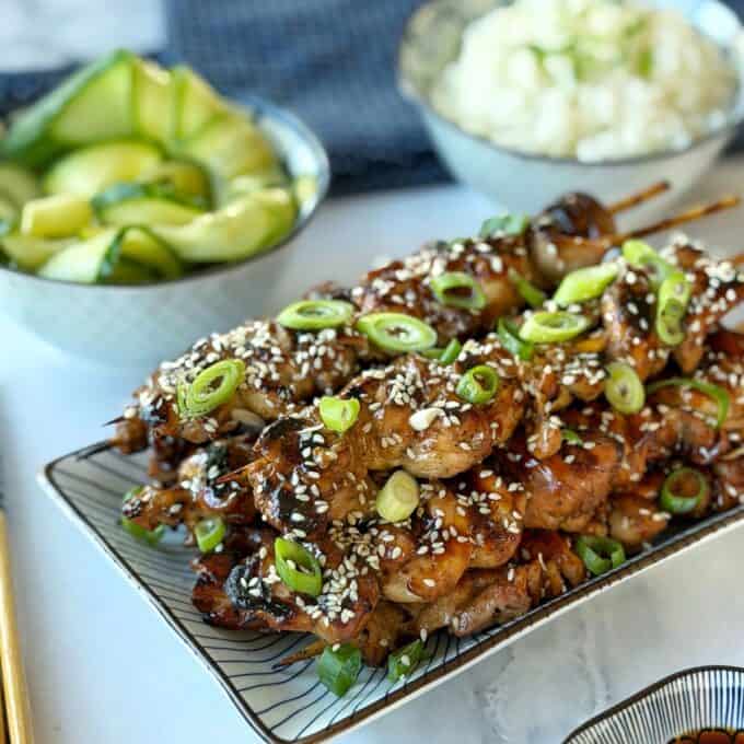 Chicken Yakitori Skewers Grilled chicken skewers topped with sesame seeds and sliced green onions, served on a rectangular plate with cucumber salad and a bowl of rice in the background.