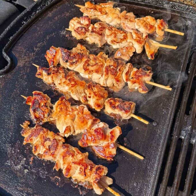 Chicken Yakitori Skewers Six grilled chicken skewers cooking on a hot, smoky grill plate.