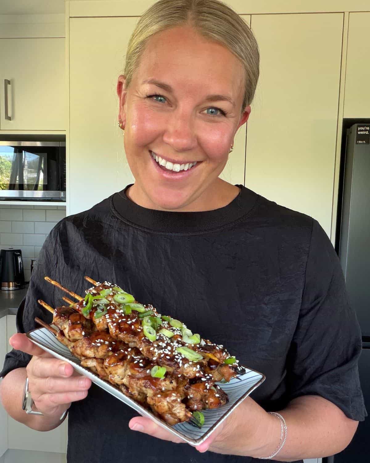 Chicken Yakitori Skewers A person smiles while holding a rectangular plate of grilled skewered meat topped with sesame seeds and sliced green onions in a kitchen.