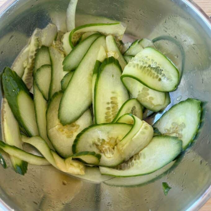 Chicken Yakitori Skewers Thinly sliced cucumber pieces in a stainless steel bowl, with visible seeds and some moisture on the vegetables and bowl.