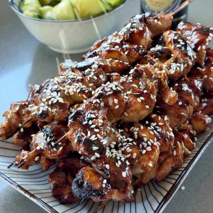 Chicken Yakitori Skewers A plate of grilled chicken skewers glazed with sauce and sprinkled with sesame seeds, with a bowl of sliced cucumbers in the background.