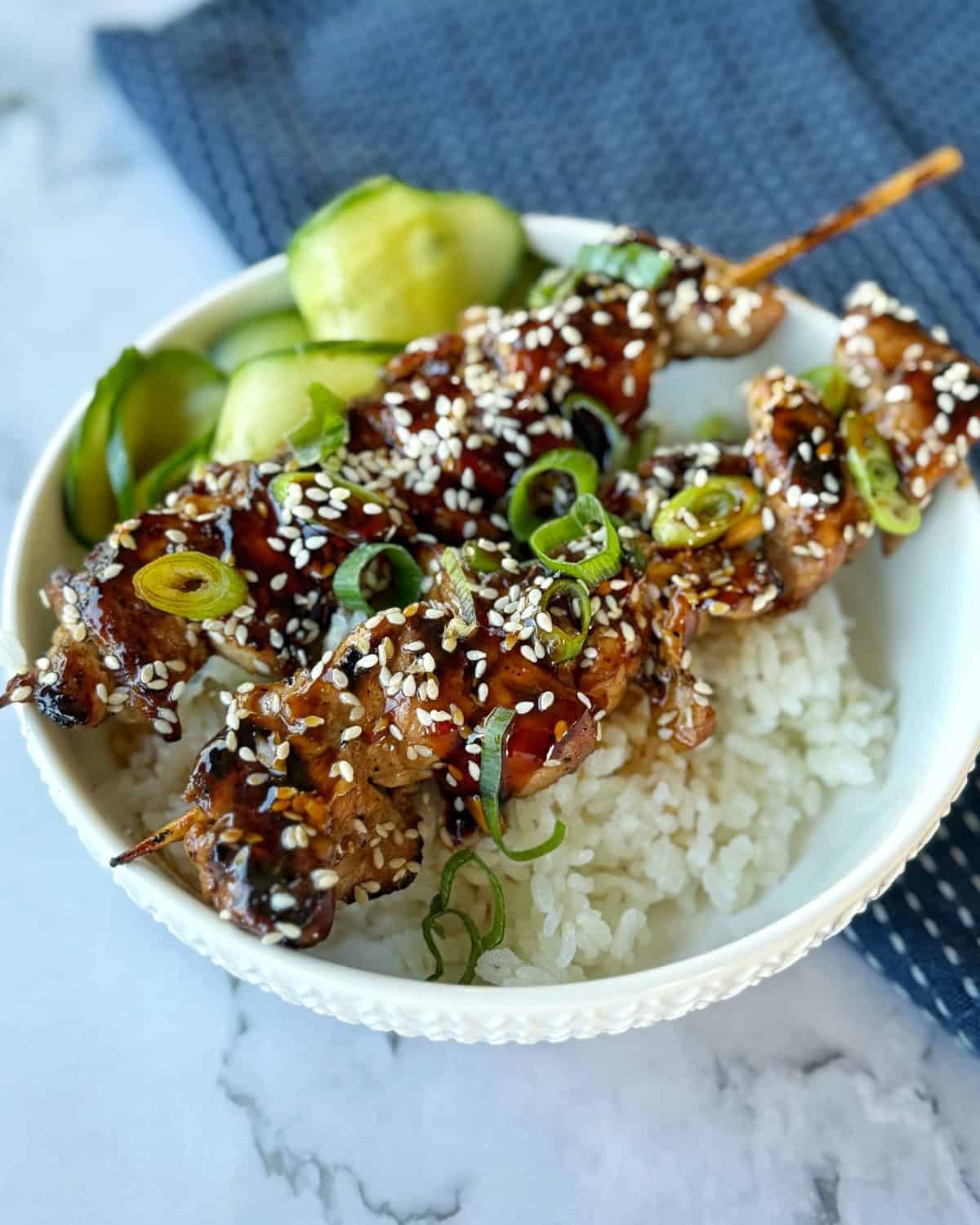 Chicken Yakitori Skewers Chicken skewers topped with sesame seeds and green onions served over white rice with sliced cucumbers in a white bowl.