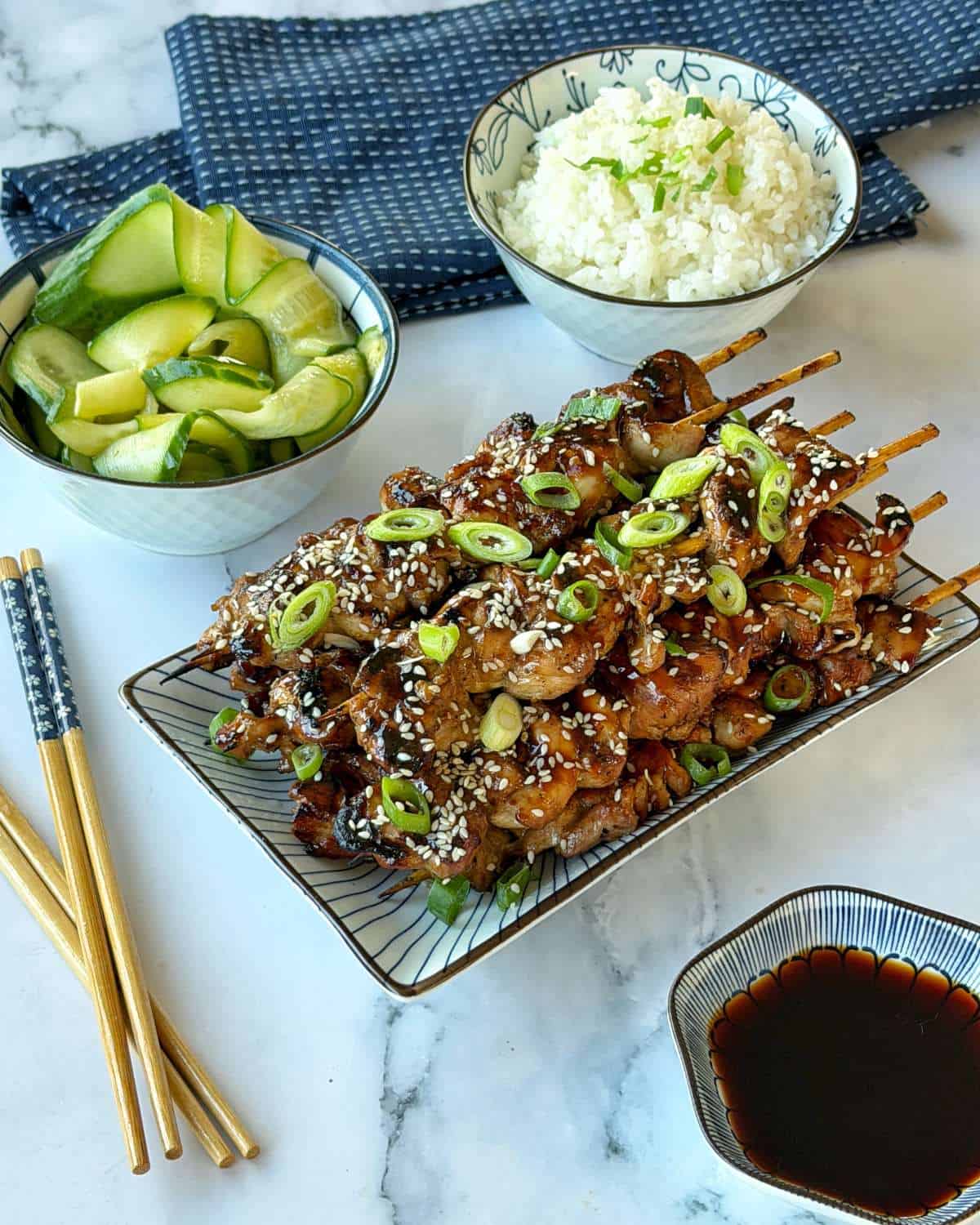 Chicken Yakitori Skewers A plate of grilled meat skewers topped with sesame seeds and scallions, served with bowls of rice, cucumber salad, and dipping sauce, with chopsticks on the side.