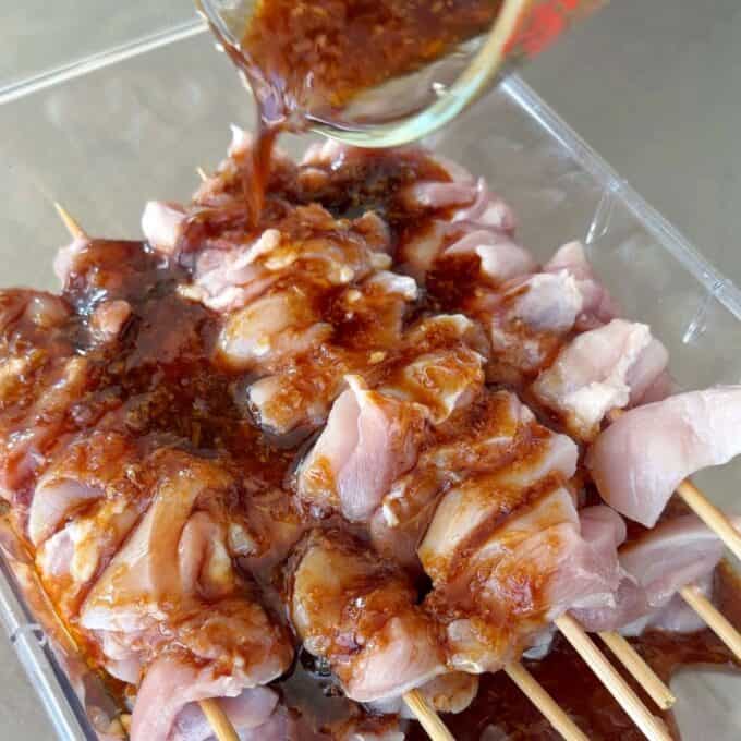 Chicken Yakitori Skewers Raw chicken skewers in a tray being covered with a dark marinade poured from a glass container.