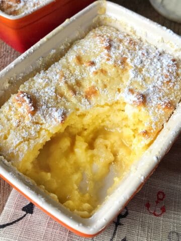 A rectangular baking dish filled with lemon pudding cake, part of the top layer is missing, revealing the soft, yellow, custard-like center. The surface is dusted with powdered sugar.