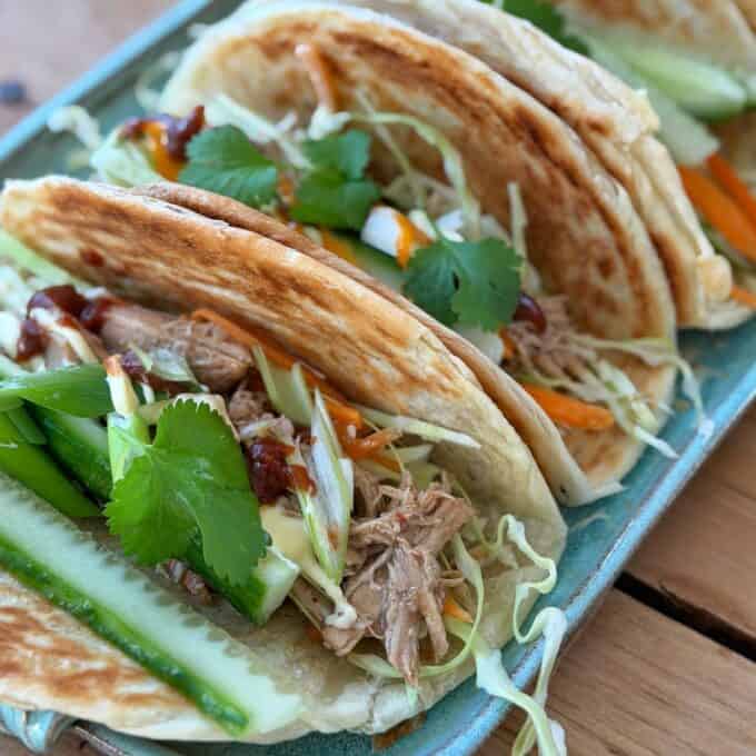 Slow Cooker Asian Style Chicken Three tacos filled with shredded meat, cucumber, green onions, shredded cabbage, carrots, cilantro, and sauce are arranged on a blue rectangular plate.