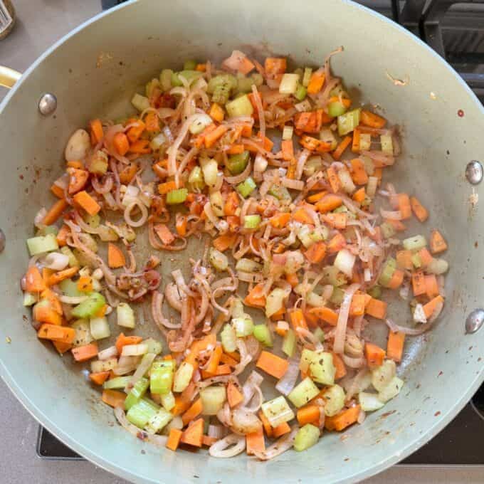 Beef Cheeks with Red Wine Sauce Chopped carrots, celery, onions, and garlic are sautéing with herbs in a large light green pan on a stove.
