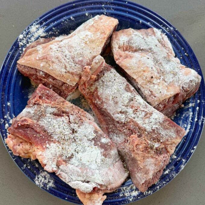 Beef Cheeks with Red Wine Sauce Four raw beef short ribs coated with flour and seasoning are arranged on a blue plate.