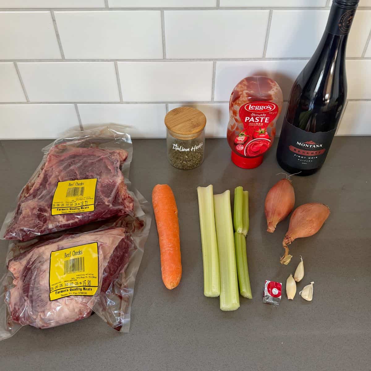 Beef Cheeks with Red Wine Sauce Two packs of beef oxtail, a jar of Italian herbs, tomato paste, red wine, a carrot, celery stalks, shallots, garlic cloves, and a beef bouillon cube on a kitchen counter.