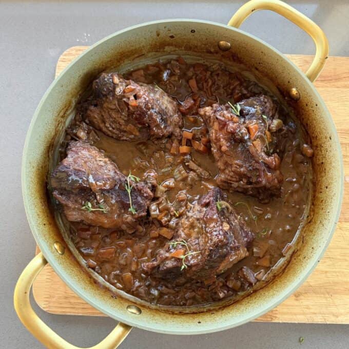 Beef Cheeks with Red Wine Sauce A round pot with four braised beef short ribs in a rich, brown sauce with chopped vegetables, garnished with sprigs of fresh herbs, sits on a wooden cutting board.