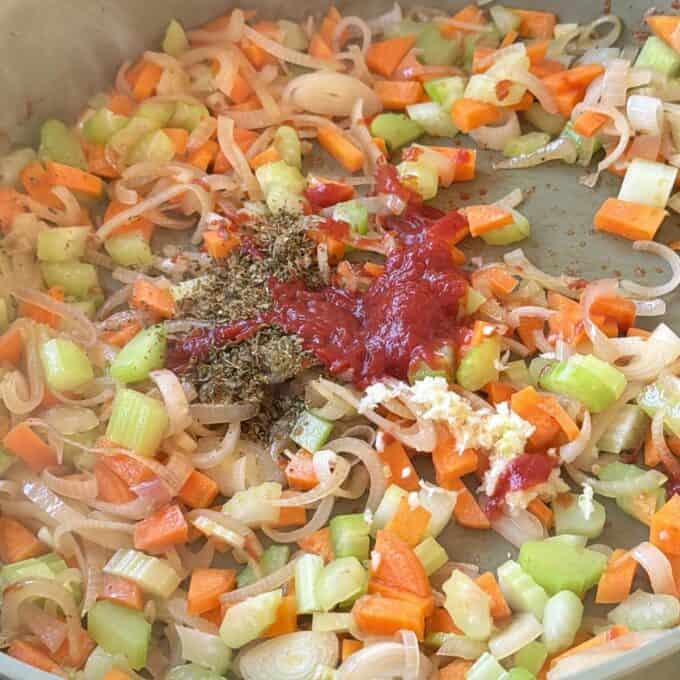 Beef Cheeks with Red Wine Sauce Chopped carrots, celery, and onions cooking in a pan with tomato paste, minced garlic, and dried herbs sprinkled on top.