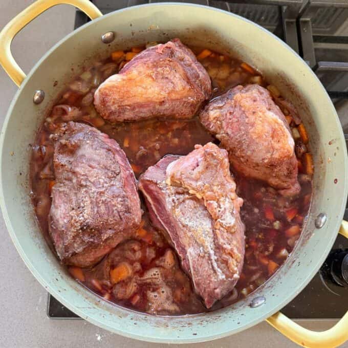 Beef Cheeks with Red Wine Sauce A pot with four large browned beef pieces simmering in a mixture of chopped vegetables and liquid on a stove.