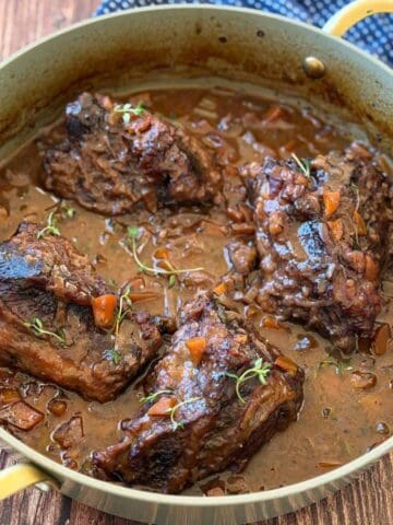 Four braised beef short ribs with vegetables and sauce in a round pan, garnished with fresh herbs, on a wooden surface.