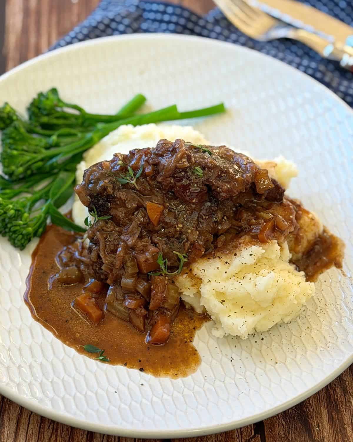 Beef Cheeks with Red Wine Sauce Braised meat with brown gravy served over mashed potatoes, garnished with herbs, and accompanied by steamed broccolini on a textured white plate.