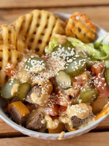 A bowl with waffle fries, lettuce, diced tomatoes, sliced pickles, beef pieces, sesame seeds, and a creamy sauce on top, placed on a wooden surface.