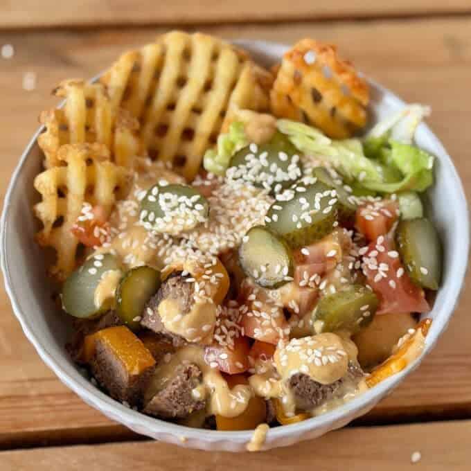 Big Mac Burger Bowls A bowl with waffle fries, lettuce, diced tomatoes, sliced pickles, beef pieces, sesame seeds, and a creamy sauce on top, placed on a wooden surface.