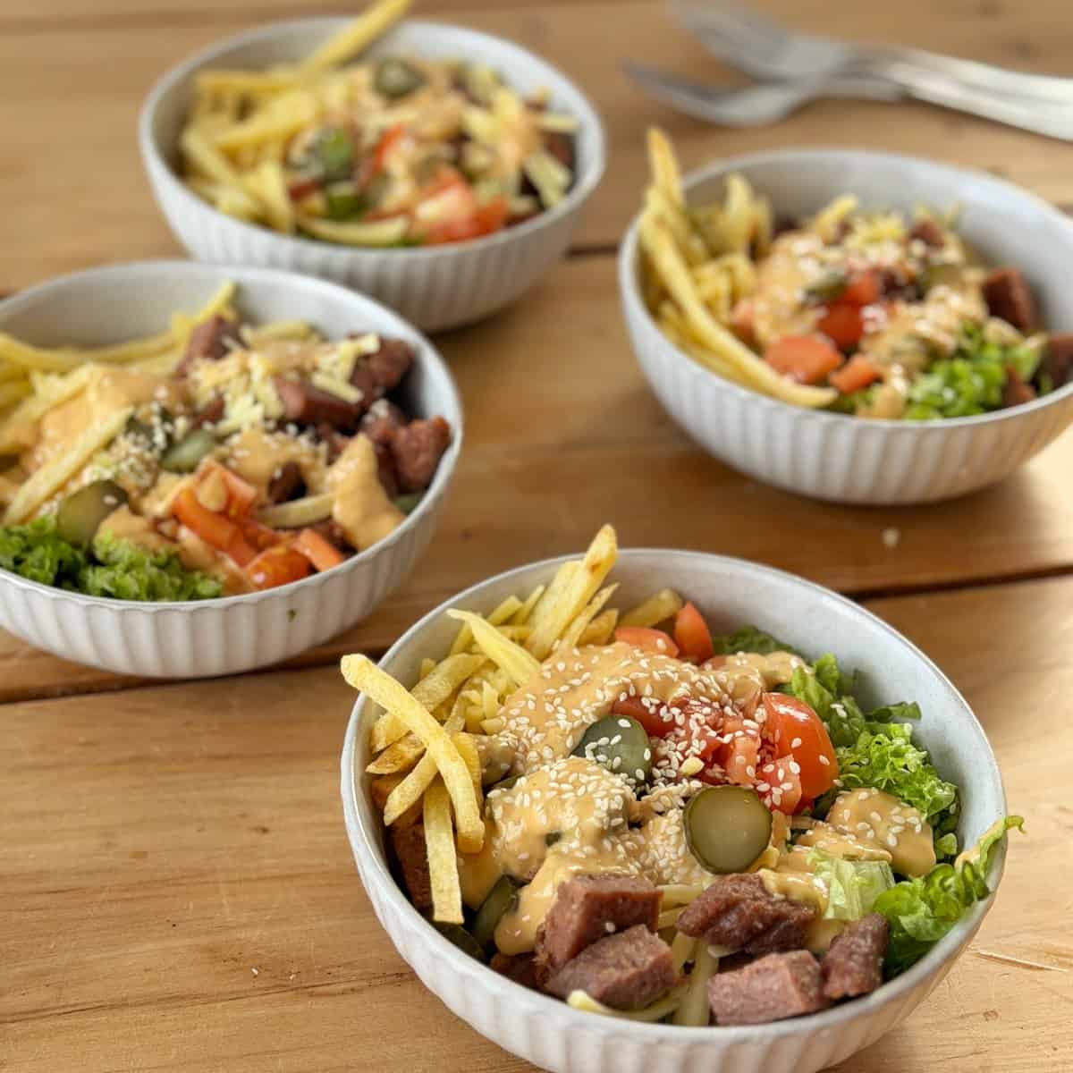 Big Mac Burger Bowls Four bowls of salad with lettuce, tomatoes, pickles, steak pieces, fries, sesame seeds, and creamy dressing are arranged on a wooden table with three metal forks in the background.