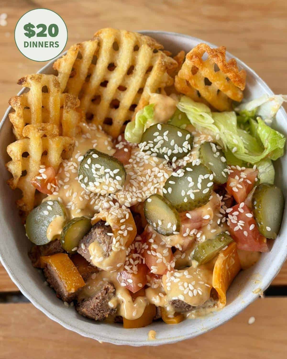 Bowl with waffle fries, lettuce, tomato, pickles, beef, sesame seeds, and creamy sauce on a wooden table. A green label reads $20 Dinners in the top left corner.