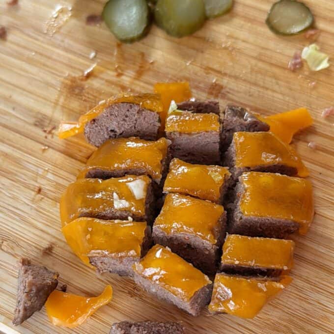 Big Mac Burger Bowls Cooked ground beef patty topped with melted cheddar cheese, cut into small squares on a wooden cutting board, with pickle slices in the background.