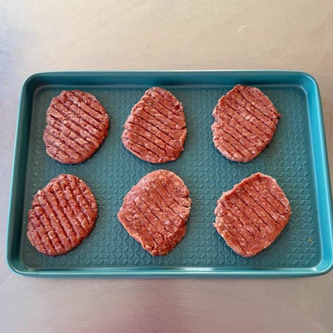 Big Mac Burger Bowls Six raw hamburger patties are arranged in two rows on a teal baking tray with a textured surface, sitting on a metal countertop.