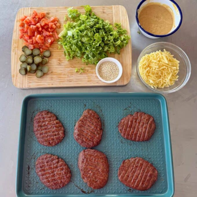 Big Mac Burger Bowls Six cooked burger patties on a tray, with chopped lettuce, tomatoes, pickles, shredded cheese, sesame seeds, and a bowl of sauce arranged on a cutting board and nearby bowls.