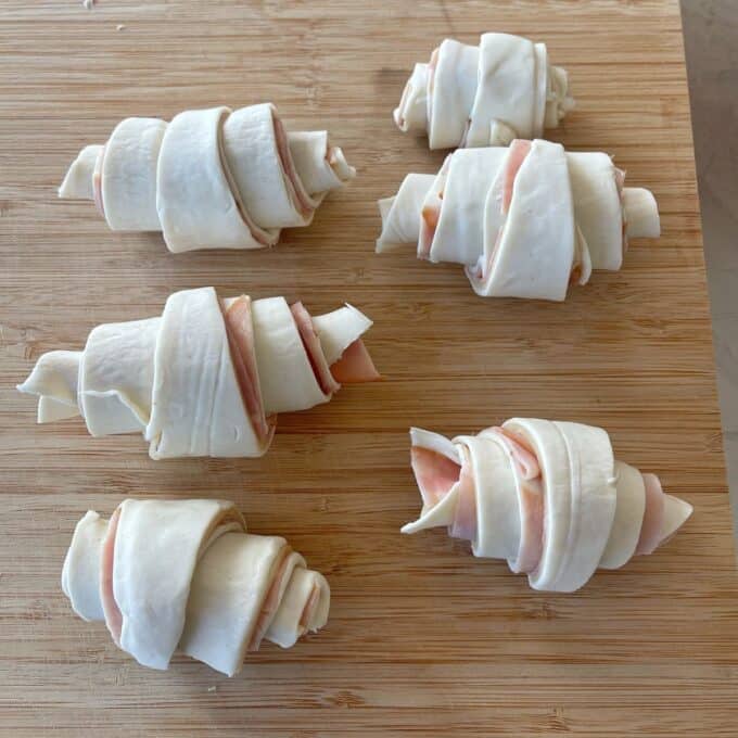 Ham and Cheese Puff Pastry Croissants Five unbaked croissants filled with ham are arranged on a wooden cutting board.