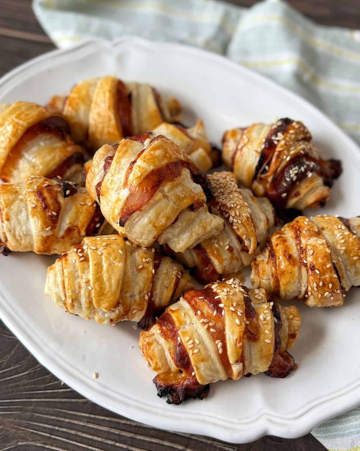 Ham and Cheese Puff Pastry Croissants A white plate holds several golden-brown puff pastry rolls, some sprinkled with sesame seeds, on a wooden surface with a striped cloth in the background.