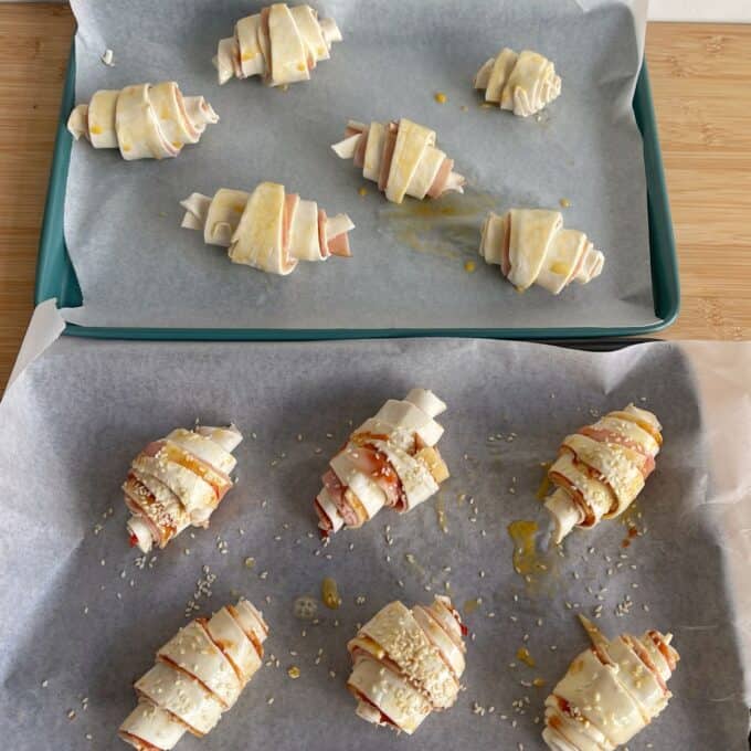 Ham and Cheese Puff Pastry Croissants Two trays of unbaked croissant rolls filled with ham and cheese, some topped with sesame seeds, arranged on parchment paper.