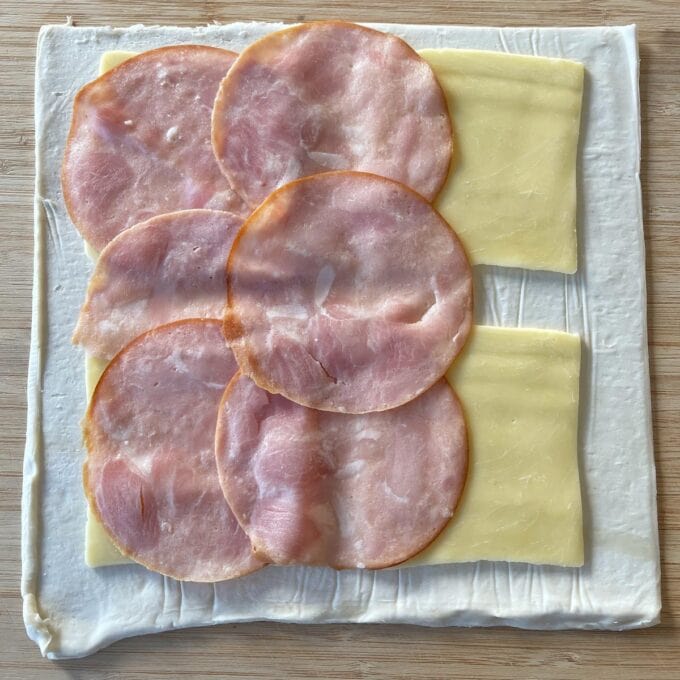 Ham and Cheese Puff Pastry Croissants A square sheet of puff pastry topped with overlapping slices of ham and rectangular slices of cheese on a wooden surface.