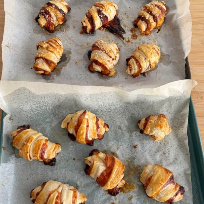 Ham and Cheese Puff Pastry Croissants Two baking trays lined with parchment paper, each holding several baked, golden-brown croissants with chocolate filling, some topped with sesame seeds.