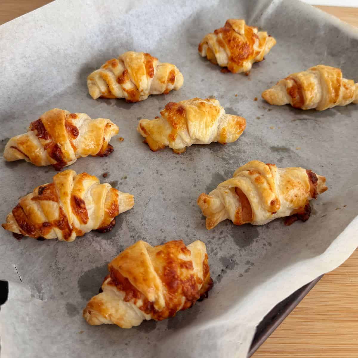 Ham and Cheese Puff Pastry Croissants Eight baked cheese croissants are arranged on a parchment-lined baking tray, with some melted cheese visible on the pastry.