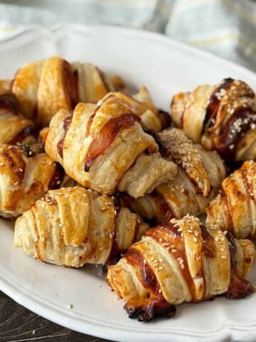 A white plate holds several golden-brown, flaky croissants with sesame seeds on top. A striped cloth is partially visible in the background.