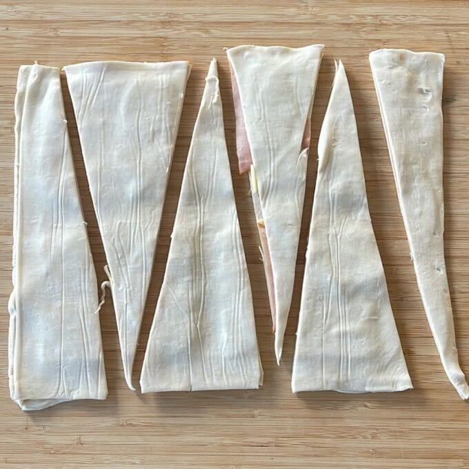 Ham and Cheese Puff Pastry Croissants Six triangular strips of dough are laid out on a wooden surface, evenly spaced, ready for further preparation.