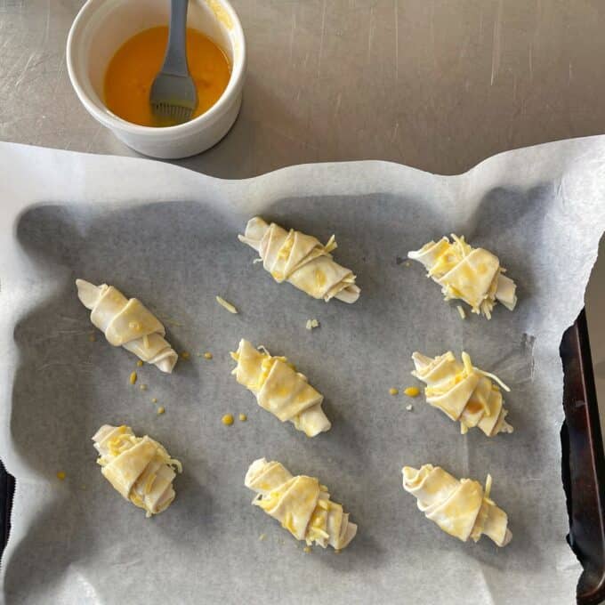 Ham and Cheese Puff Pastry Croissants Eight unbaked pastry rolls on a parchment-lined tray, brushed with egg wash, with a bowl of egg mixture and brush nearby on a metal surface.