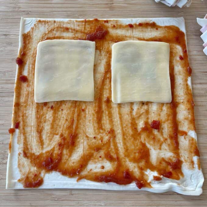 Ham and Cheese Puff Pastry Croissants A sheet of dough spread with tomato sauce, topped with two slices of cheese, sits on a wooden surface.