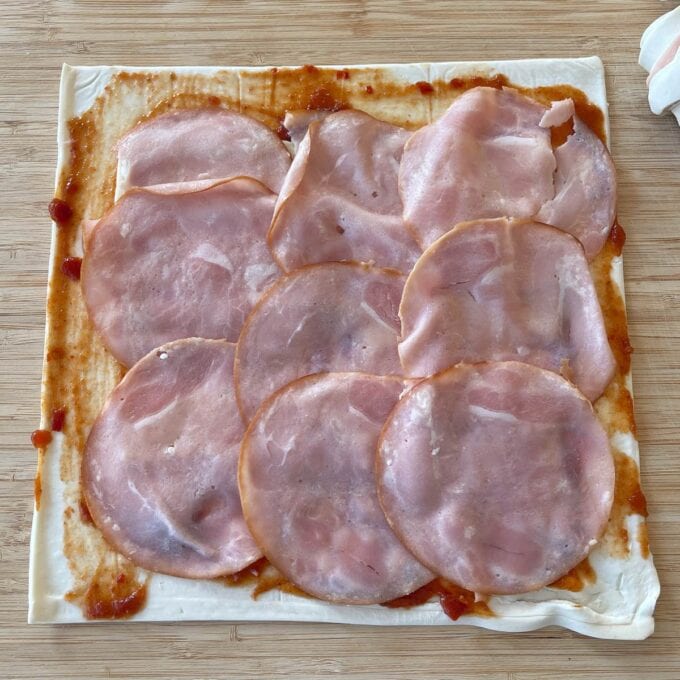 Ham and Cheese Puff Pastry Croissants A square sheet of pastry covered with tomato sauce, topped with overlapping slices of ham, on a wooden surface.