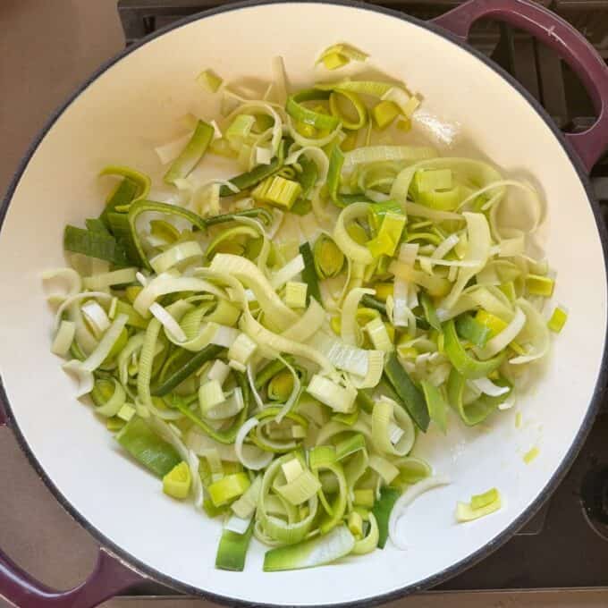 Potato Leek Gratin Sliced leeks cooking in a white pan on a stove, with green and white pieces spread out evenly.