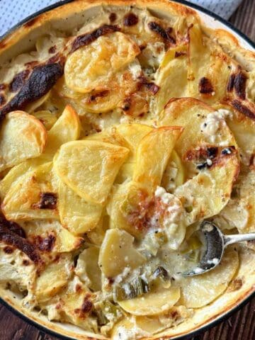 A baked dish of scalloped potatoes in a creamy sauce sits in a round pan, with a serving spoon partially submerged in the casserole.
