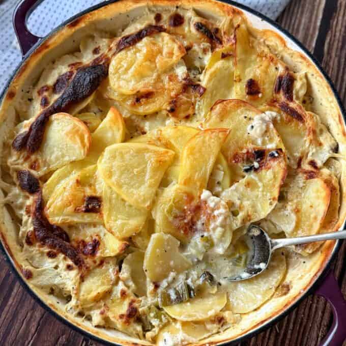 Potato Leek Gratin A baked dish of scalloped potatoes in a creamy sauce sits in a round pan, with a serving spoon partially submerged in the casserole.