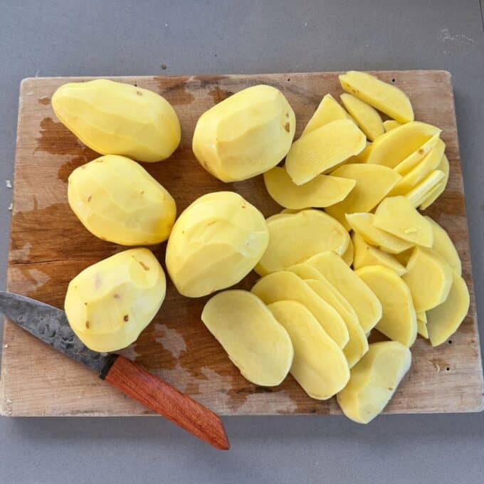 Potato Leek Gratin Peeled whole and sliced potatoes are arranged on a wooden cutting board next to a knife with a wooden handle.