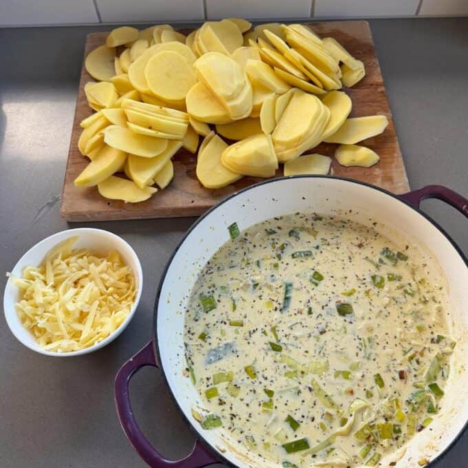 Potato Leek Gratin Sliced potatoes on a board, grated cheese in a bowl, and a creamy herb mixture with leeks in a casserole dish on a kitchen counter.